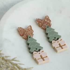 May include: A pair of Christmas-themed earrings featuring a pink bow, a green layered Christmas tree, and a pink gift box.