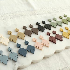 May include: A collection of leaf-shaped earrings in various colors, including cream, tan, blue, pink, brown, green, yellow, and purple. The earrings are displayed on a white, scalloped tray.