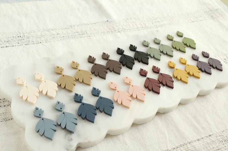 May include: A collection of leaf-shaped earrings in various colors, including cream, tan, blue, pink, brown, green, yellow, and purple. The earrings are displayed on a white, scalloped tray.