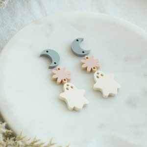 May include: Two pairs of Halloween-themed earrings with a moon, flower, and ghost charm in shades of gray, pink, and white.