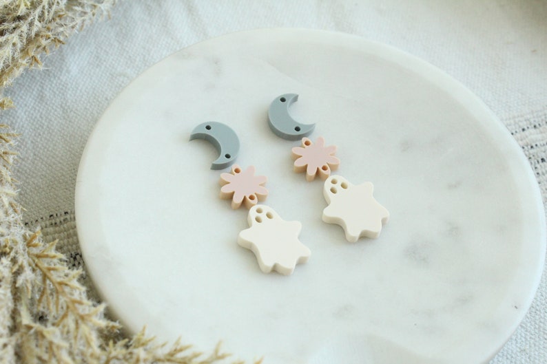 May include: Two pairs of Halloween-themed earrings with a moon, flower, and ghost charm in shades of gray, pink, and white.