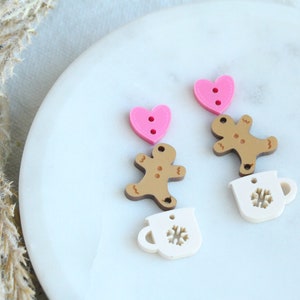 May include: A collection of decorative buttons in various shapes. The image features pink heart-shaped buttons, gingerbread man buttons, and white mug-shaped buttons with snowflake cutouts. The buttons are arranged on a white surface.