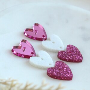 May include: Four pink and white heart-shaped embellishments. Two hearts are solid pink, one is white with a pink glitter overlay, and one is pink with a white glitter overlay.