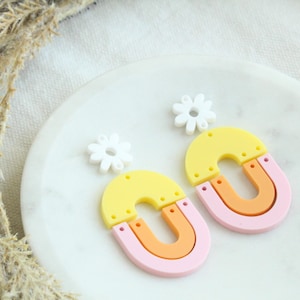 May include: A pair of colorful statement earrings featuring a yellow, orange, and pink layered arch design with a white daisy charm.
