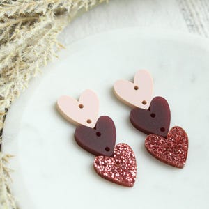 May include: A pair of earrings with three layered heart charms. The top heart is pink, the middle heart is burgundy, and the bottom heart is a sparkly pink.