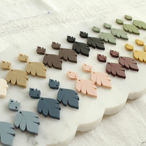 May include: A collection of leaf-shaped embellishments in various colors, including cream, tan, brown, pink, blue, green, and yellow. The leaves are arranged on a white, scalloped-edge surface, suggesting a craft or decorative theme.