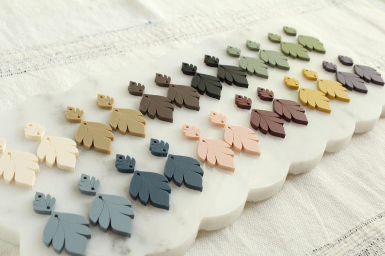 May include: A collection of leaf-shaped embellishments in various colors, including cream, tan, brown, pink, blue, green, and yellow. The leaves are arranged on a white, scalloped-edge surface, suggesting a craft or decorative theme.