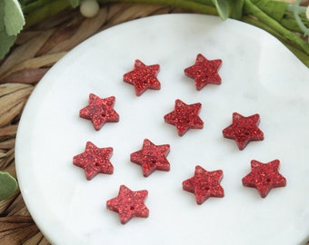 May include: Ten red glitter star-shaped embellishments on a white surface.