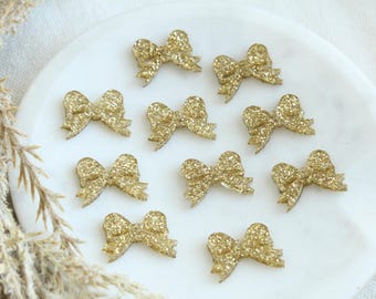 May include: Ten gold glitter bows on a white surface. The bows are all the same size and shape, and they are arranged in a random pattern.