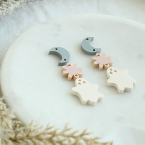 May include: A pair of dangle earrings with a moon, flower, and ghost charm. The charms are made of white, light pink, and gray polymer clay.