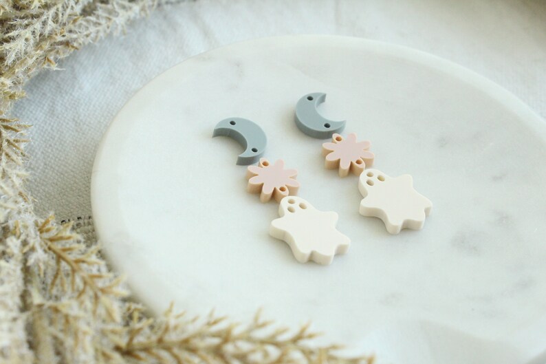 May include: A pair of dangle earrings with a moon, flower, and ghost charm. The charms are made of white, light pink, and gray polymer clay.