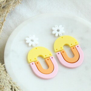 May include: A pair of colorful statement earrings featuring a yellow, orange, and pink layered design with a white daisy charm.