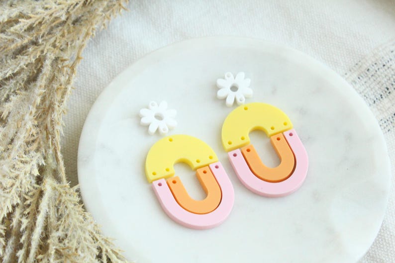 May include: A pair of colorful statement earrings featuring a yellow, orange, and pink layered design with a white daisy charm.