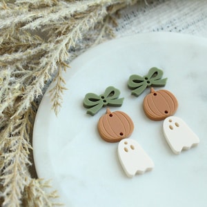 May include: A collection of fall-themed embellishments, including olive green bows, brown pumpkins, and off-white ghost shapes. These small, decorative pieces are arranged on a white, oval-shaped surface, perfect for crafting projects.