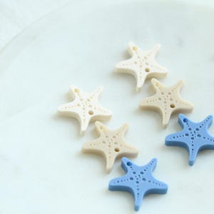 May include: A collection of starfish-shaped buttons in various colors, including off-white, tan, and light blue. The buttons are arranged on a white marble surface. Each button has a small hole for sewing.