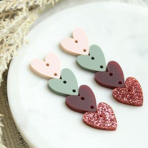 May include: A pair of dangle earrings with a layered heart design. The hearts are pink, green, burgundy, and sparkly pink.