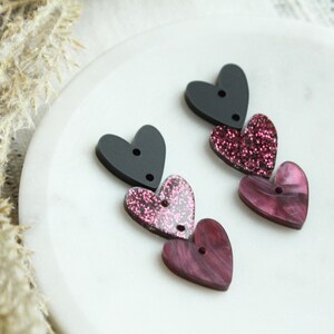May include: Three heart-shaped charms in black, pink glitter, and marbled pink. The charms are strung together and have two holes for attaching to a necklace or bracelet.