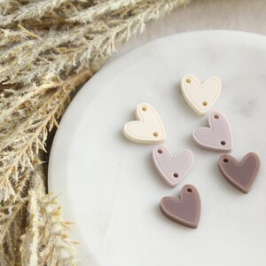 May include: A collection of heart-shaped silicone beads in various muted colors: cream, lavender, and mauve. Each bead has two small holes for stringing. The beads are arranged on a white, marble-like surface.