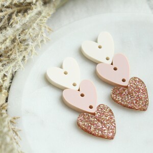 May include: A pair of dangle earrings with three layered hearts. The top heart is white, the middle heart is pink, and the bottom heart is pink with gold glitter.