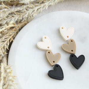 May include: Six heart-shaped silicone beads in cream, tan, and black. Each bead has two small holes for stringing. The beads are arranged on a white marble surface, with dried ornamental grass in the background.