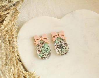 May include: Two decorative earrings with a floral pattern in shades of green, pink, and white. Each earring has a light pink bow at the top. The earrings are egg-shaped and sit on a white, heart-shaped surface.