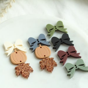 May include: A collection of fall-themed embellishments, including bow shapes, pumpkins, and maple leaves. The embellishments are in various colors, such as cream, blue, green, black, and burgundy. The maple leaves have a glittery finish.