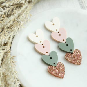 May include: A pair of dangle earrings with a cascade of three heart-shaped charms. The hearts are in shades of pink, white, and green, with the bottom heart featuring a sparkly gold finish.