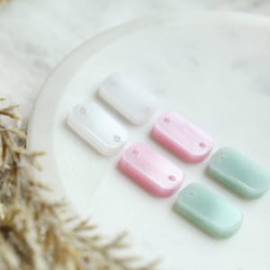 May include: Six rectangular resin charms in various pastel colors, including white, pink, and green. Each charm has two small holes for attaching to jewelry or other crafts.