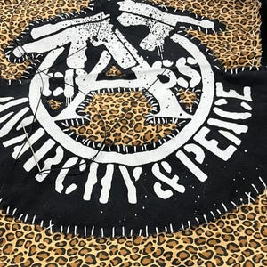 Puede incluir: Parche negro con texto blanco "ANARCHY & PEACE" cosido sobre una tela con estampado de leopardo. El parche presenta un diseño circular con un gráfico central y está cosido con hilo blanco.