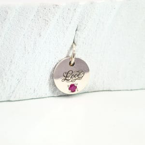 May include: A round silver pendant with the word "Love" in cursive script. A small, red gemstone is set at the bottom. The pendant hangs from a small loop at the top.