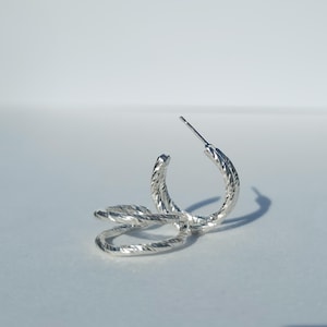 May include: Silver hoop earrings with a twisted design. The earrings are made of a textured metal and have a simple, modern style.