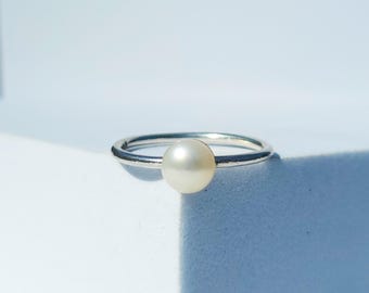 May include: A minimalist silver ring featuring a single, round, cream-colored pearl. The ring's band is thin and polished, and the pearl is centered, creating a simple yet elegant design. The ring is set against a bright, white background.