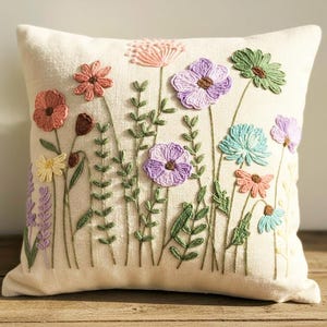 May include: Cream-colored square throw pillow with embroidered flowers in various colors. The flowers include pink, purple, green, and blue, with green stems and leaves. The pillow is made of a textured fabric.