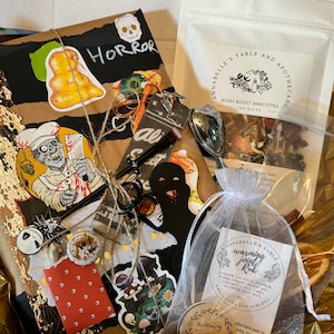 May include: A brown paper package wrapped with twine and decorated with Halloween-themed stickers. The package includes a tea blend labeled "Berry Boost Immunitea" and a small jar of "Warming Joint Rub" with a label listing ingredients like ginger, cayenne pepper, and olive oil.