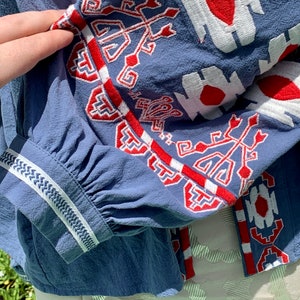 May include: A blue cotton blouse with white and red embroidered geometric patterns on the sleeves and neckline.