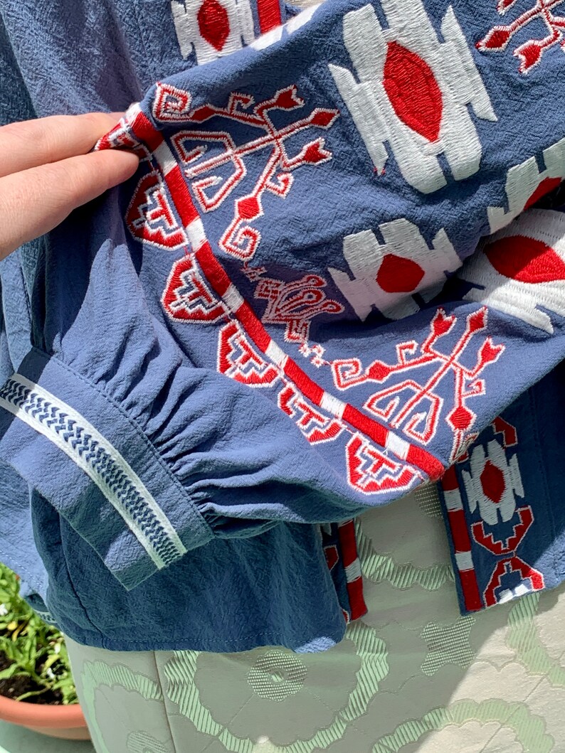 May include: A blue cotton blouse with white and red embroidered geometric patterns on the sleeves and neckline.