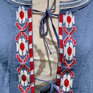 May include: A blue cotton tunic with a white and red embroidered pattern. The tunic has a tie closure at the neckline.