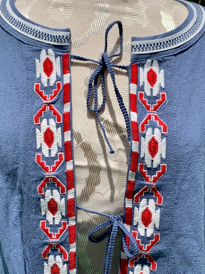 May include: A blue cotton tunic with a white and red embroidered pattern. The tunic has a tie closure at the neckline.