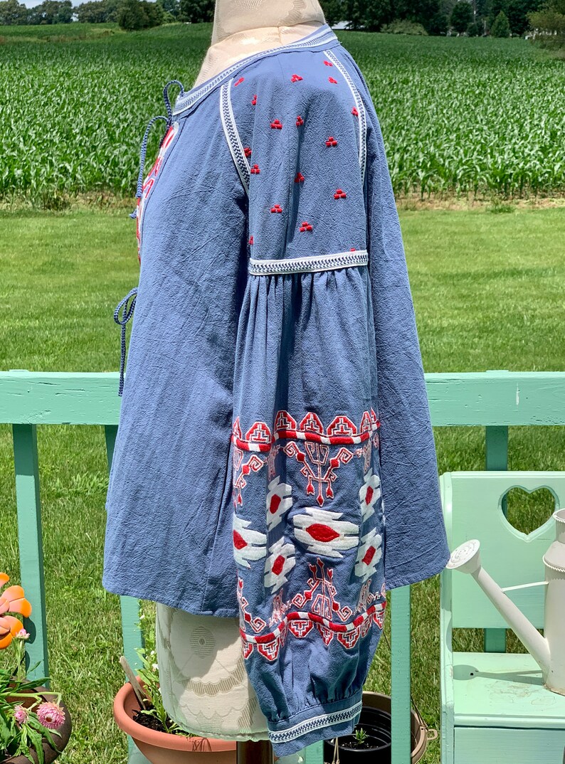 May include: A blue cotton blouse with red and white embroidery. The blouse has a tie at the neck and long sleeves with a gathered cuff. The embroidery is a geometric pattern.