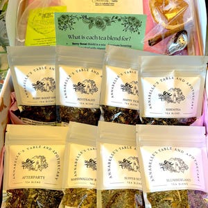 May include: A collection of eight small, white, resealable pouches filled with loose leaf tea blends. Each pouch has a label with the name of the tea blend, "Annabelle's Table and Apothecary", and a description of the ingredients. The tea blends are: Berry Boost Shield, Revitalize, Happy Tick, Serenitea, Afterparty, Marshmallow Root, Supper Set, and Slumberland.