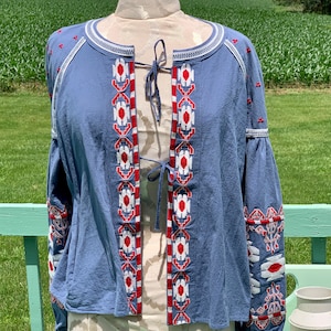 May include: A blue cotton jacket with white and red embroidered geometric patterns. The jacket has a tie closure at the neckline and long sleeves.