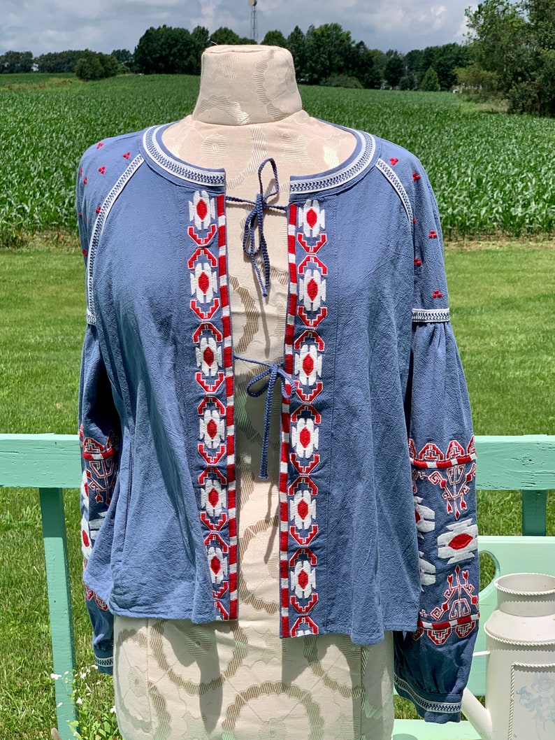 May include: A blue cotton jacket with white and red embroidered geometric patterns. The jacket has a tie closure at the neckline and long sleeves.
