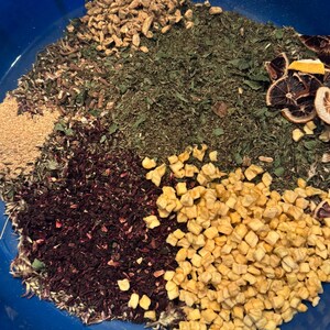 May include: A close-up of a blue bowl filled with various dried ingredients. The ingredients include dried ginger, green herbs, dried hibiscus, diced apples, and dried citrus slices. The ingredients are arranged in sections.