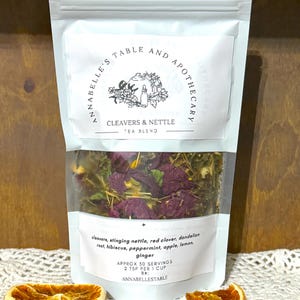 May include: A clear pouch of Cleavers & Nettle tea blend with visible herbs and flowers. The packaging includes the text "ANNABELLE'S TABLE AND APOTHECARY" and a list of ingredients. Two dried orange slices are in the foreground.