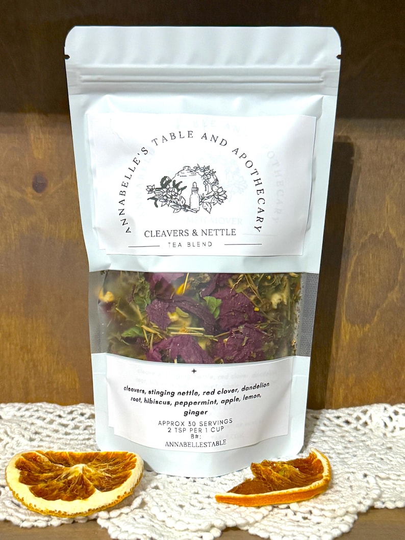 May include: A clear pouch of Cleavers & Nettle tea blend with visible herbs and flowers. The packaging includes the text "ANNABELLE'S TABLE AND APOTHECARY" and a list of ingredients. Two dried orange slices are in the foreground.