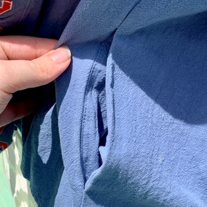 May include: Close-up of a blue fabric pocket on a garment. The pocket is sewn with a straight stitch and has a slight curve at the top.