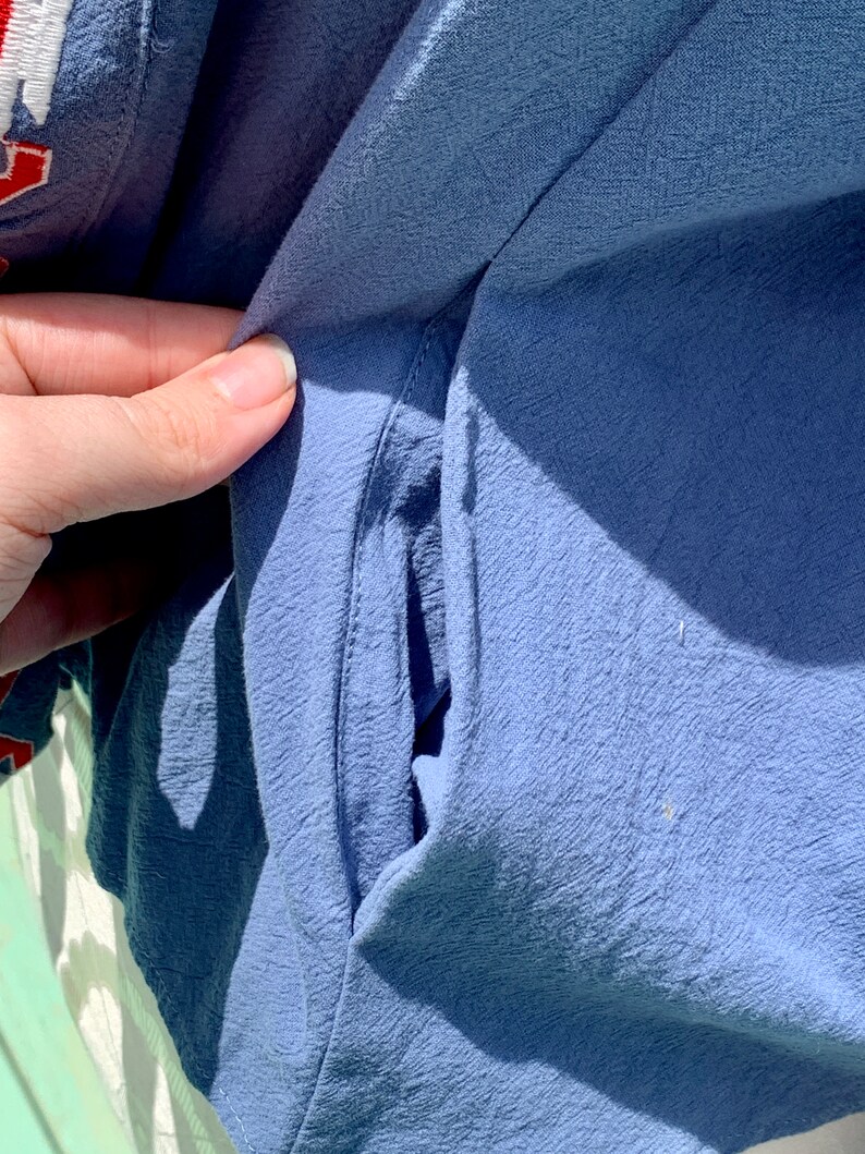 May include: Close-up of a blue fabric pocket on a garment. The pocket is sewn with a straight stitch and has a slight curve at the top.