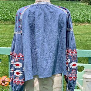 May include: A light blue blouse with long sleeves and white and red embroidered geometric patterns. The blouse has a gathered neckline and a relaxed fit.