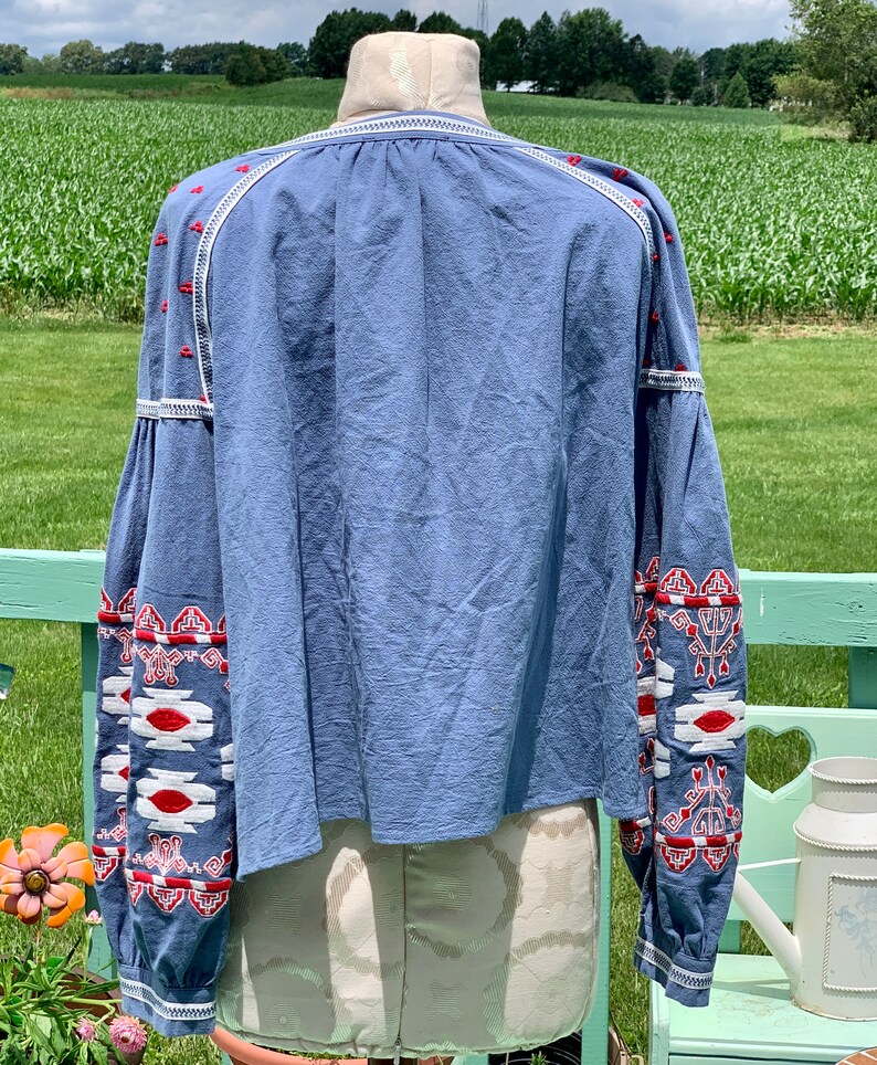 May include: A light blue blouse with long sleeves and white and red embroidered geometric patterns. The blouse has a gathered neckline and a relaxed fit.