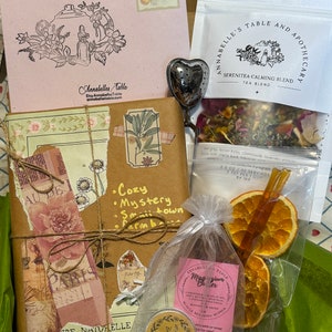 May include: A brown paper-wrapped journal with a pink floral design and a handwritten title "Cozy Mystery Small Town Farmhouse". A small bag of tea with the text "Serenitea Calming Blend Tea Blend" is in the background. A small jar of magnesium butter with the text "Annabelles Table Magnesium Butter" is in the foreground.